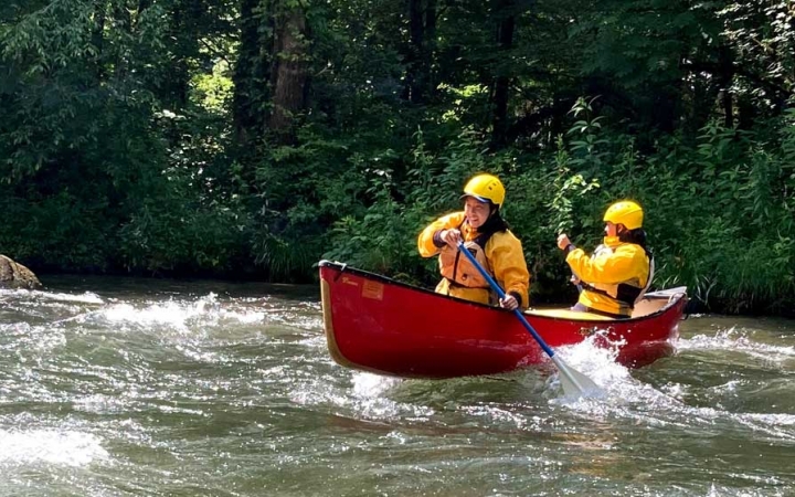 Two people wearing safety gear paddle a canoe beside a shore with thick greenery 